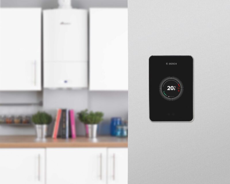 A black Bosch EasyControl smart thermostat mounted on a wall showing 20 degrees, with a blurred kitchen and wall-mounted boiler in the background