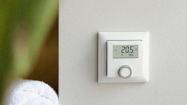 A white wall-mounted room thermostat displaying 20.5°C and 48% humidity, mounted on a wall beside a towel
