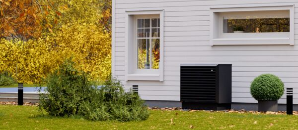 A black air-to-water heat pump unit installed outside a white weatherboard house with autumn foliage in the garden