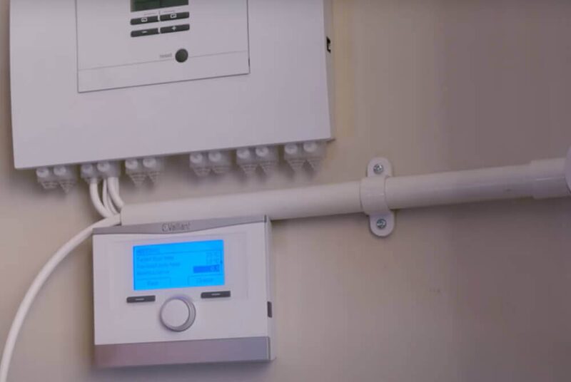 A Vaillant boiler control thermostat with blue display mounted on a wall below a wiring box and white pipework