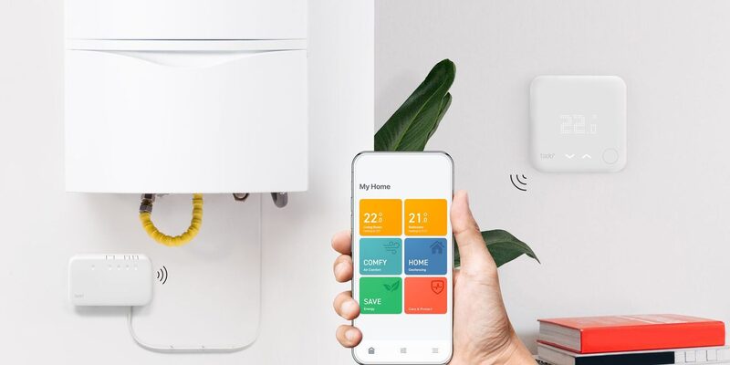 A hand holding a smartphone with a tado smart thermostat app next to a white wall-mounted tado thermostat and a boiler with a tado receiver