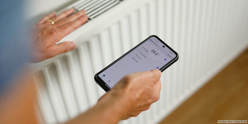 A person holding a smartphone showing a heating control app reading 23.2°C next to a white radiator