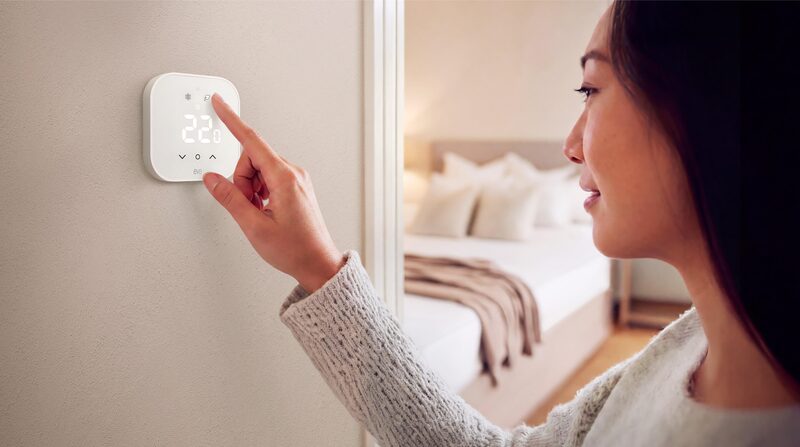 A woman adjusting a white Eve smart thermostat mounted on a bedroom wall