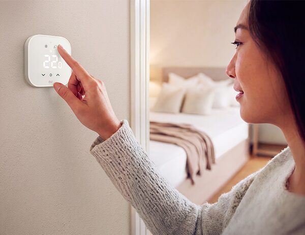 A woman touching a white Eve smart thermostat on a wall showing 22 degrees, with a bedroom in the background