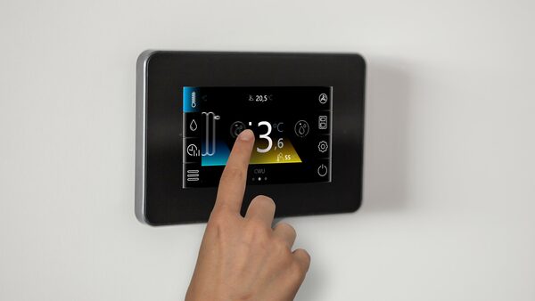 A hand touching a wall-mounted Grant Aerona smart heat pump controller with a colour touchscreen showing temperatures