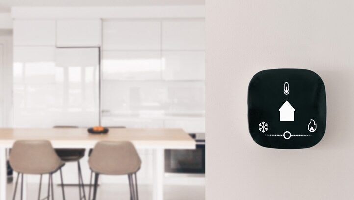 A black smart thermostat mounted on a wall beside a blurred white kitchen and dining area