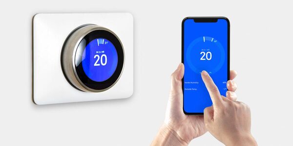 A Nest smart thermostat on a wall next to a hand controlling it via a smartphone app, both showing 20 degrees