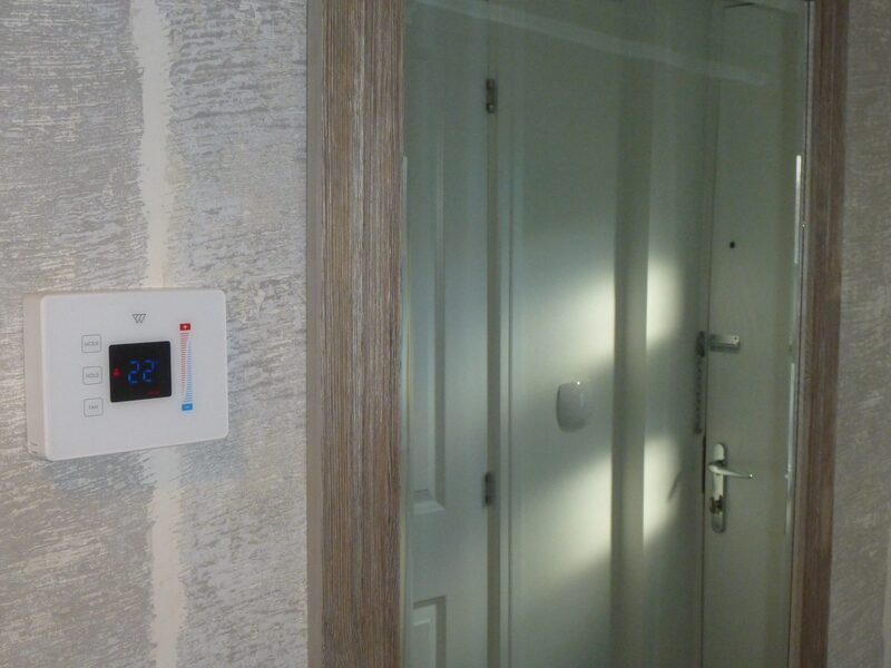 A white Wondrwall smart thermostat on a wall reading 22 degrees, next to a frosted glass door in a hallway
