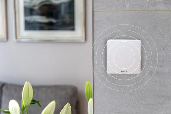 A white Wunda screenless smart room thermostat mounted on a grey tiled wall with illustrated signal rings, in a home interior