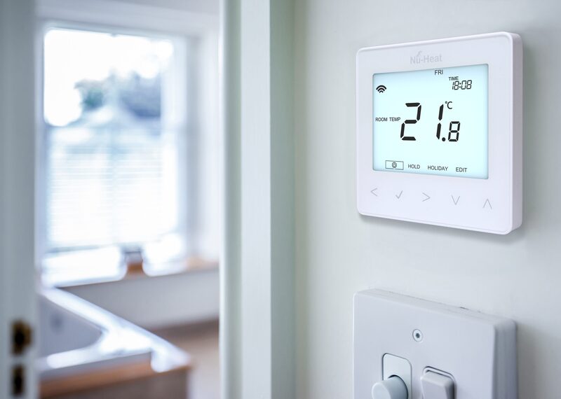 A white Nu-Heat smart thermostat mounted on a wall showing a room temperature of 21.8°C, with a light switch below and a blurred bathroom in the background