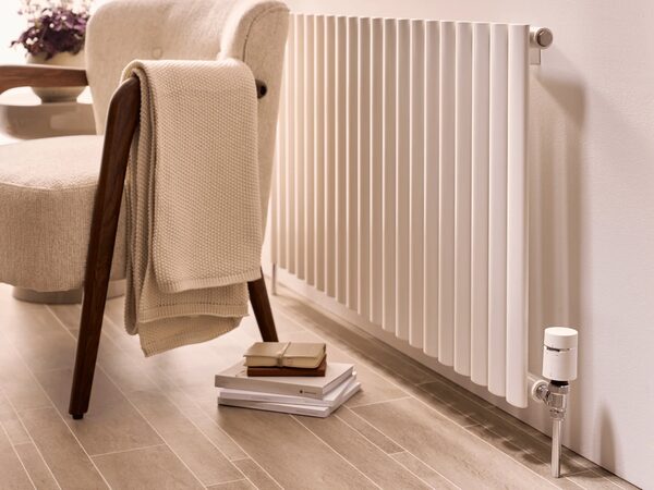 A white vertical column radiator with a smart thermostatic radiator valve fitted, next to an armchair in a living room