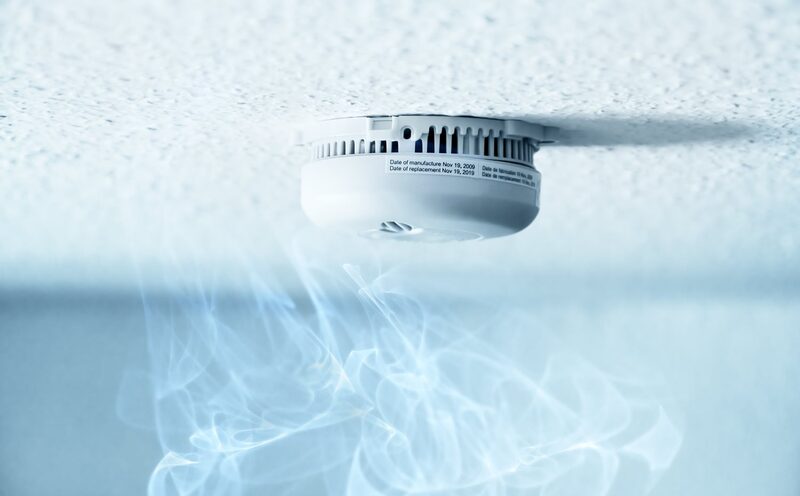 A white ceiling-mounted smoke alarm with wisps of smoke rising towards it