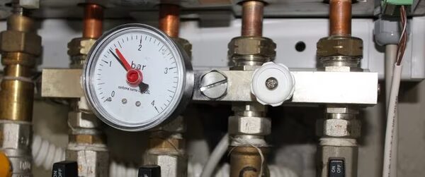 A close-up of a boiler pressure gauge reading in bar, mounted among brass valves and copper pipework on a heating manifold