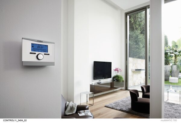 A Vaillant multiMATIC 700 wall-mounted heating control thermostat with blue display on a white wall in a modern living room