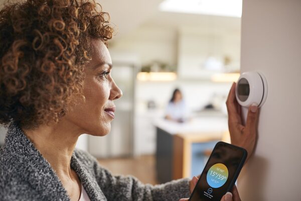 A woman adjusting a wall-mounted smart thermostat while holding a smartphone showing the matching heating control app