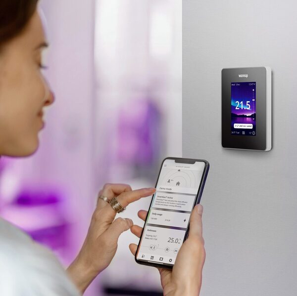 A woman using the Warmup app on her phone next to a Warmup smart thermostat mounted on a wall