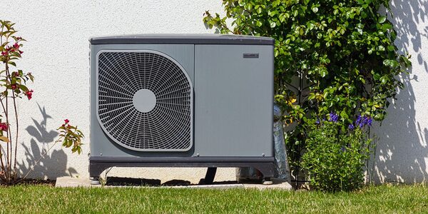 An air source heat pump unit installed outside a house against a white wall, surrounded by garden plants