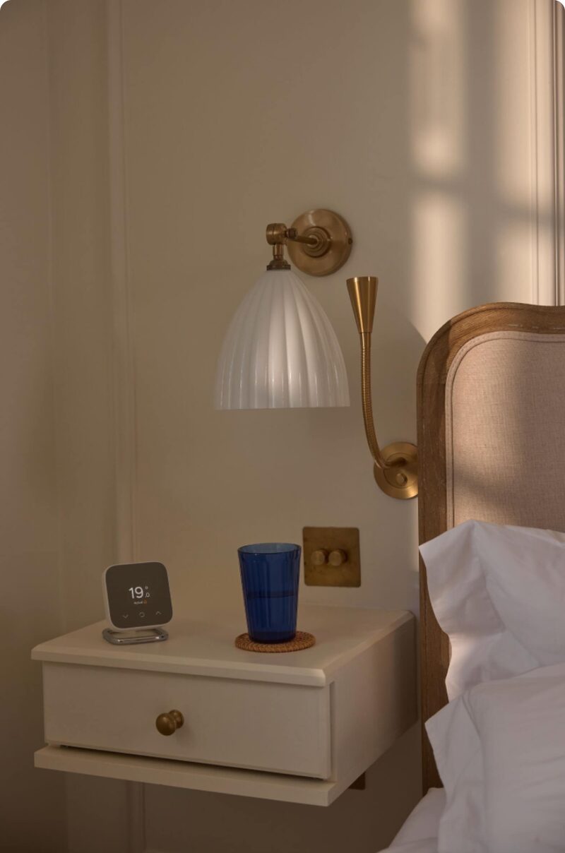 A Hive smart thermostat on a stand sitting on a bedside table next to a bed and wall lamp
