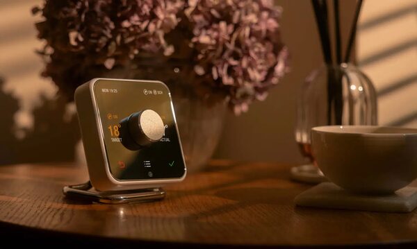 A Hive smart thermostat on a metal stand on a wooden table showing 18 degrees, with flowers and a cup in the background