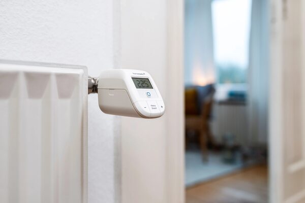 A white Homematic IP smart radiator thermostat fitted on a radiator, showing 22.5 degrees, with a blurred home interior in the background.