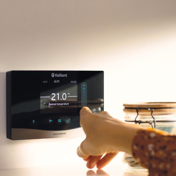A hand adjusting a wall-mounted Vaillant sensoCOMFORT heating thermostat set to 21.0°C