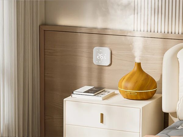 A Meross smart thermostat displaying 26.5°C mounted on a wooden bedroom wall above a bedside table with a diffuser