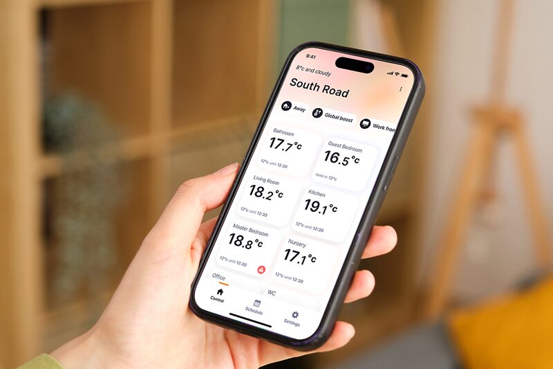 A hand holding a smartphone displaying the Heatmiser Neo app with room-by-room heating temperatures