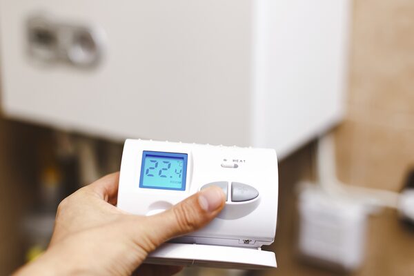 A hand holding a white digital room thermostat showing 22.4°C in front of a wall-mounted boiler