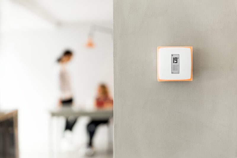 A Netatmo smart thermostat mounted on a wall showing 19 degrees, with a blurred family in the background
