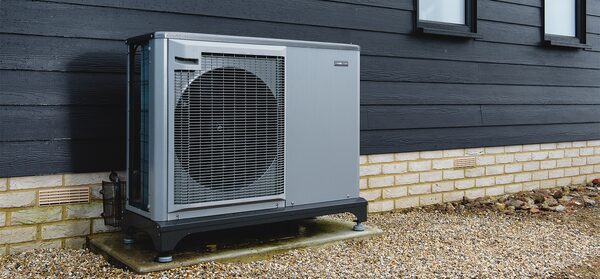 A grey air source heat pump unit installed outside a house with dark cladding and a brick base