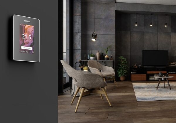 A Warmup smart thermostat mounted on a dark wall beside a modern living room with armchairs and a TV