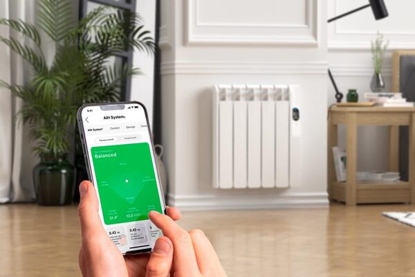 A hand holding a smartphone showing a heating control app in front of a white Rointe electric radiator installed in a living room