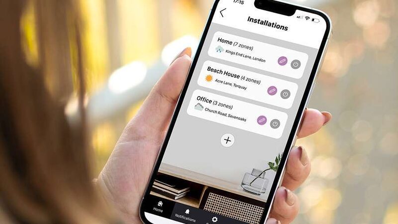 A hand holding a smartphone showing the Rointe app Installations screen listing heating zones for Home, Beach House and Office