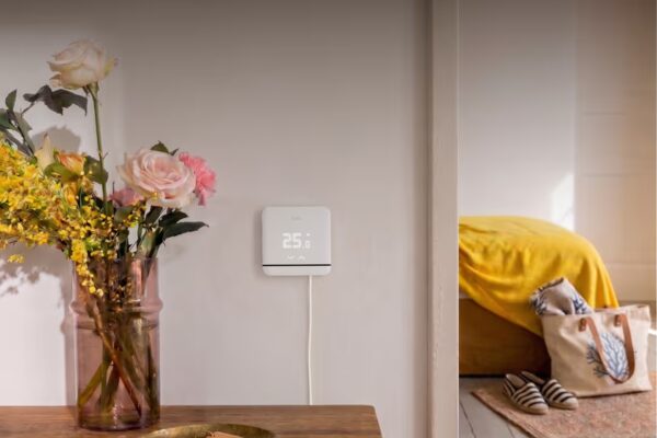 A white tado smart thermostat mounted on a wall showing 25.0 degrees, next to a vase of flowers in a home interior