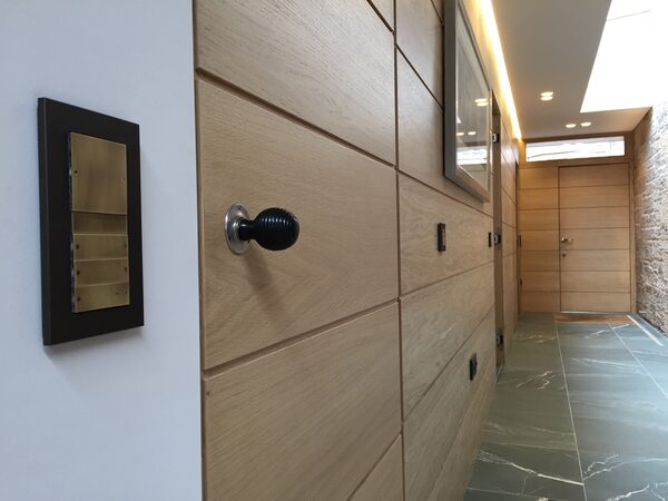 A brass KNX wall control panel beside a wooden panelled door in a modern home hallway