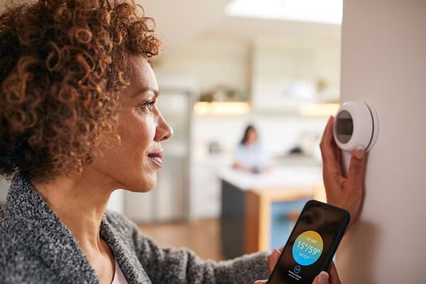 A woman adjusting a white smart thermostat on a wall while holding a smartphone showing the temperature