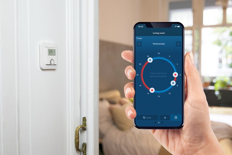 A hand holding a smartphone showing a thermostat timer app next to a wall-mounted room thermostat in a home