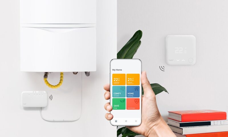 A hand holding a smartphone with a tado smart thermostat app next to a white wall-mounted tado thermostat and a boiler with a tado receiver
