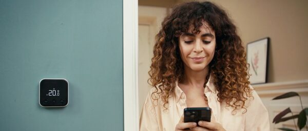 A tado° smart thermostat on a teal wall showing 20°C beside a woman checking her phone app in a home