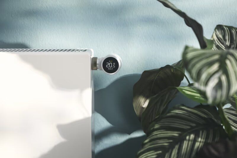 A tado° Smart Radiator Thermostat X mounted on a white radiator, its round display reading 20.5°C, beside a houseplant against a blue wall
