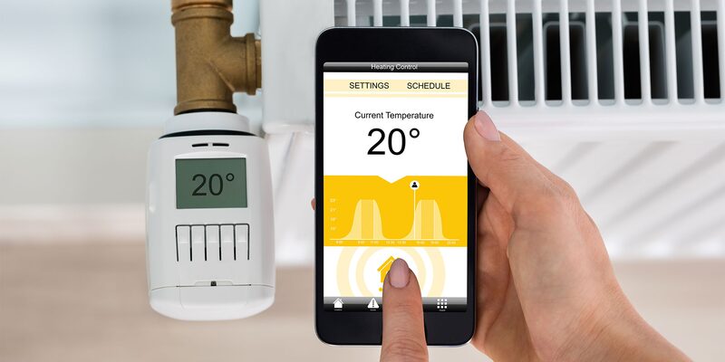 A hand holding a smartphone showing a heating control app set to 20 degrees next to a smart radiator valve fitted on a radiator