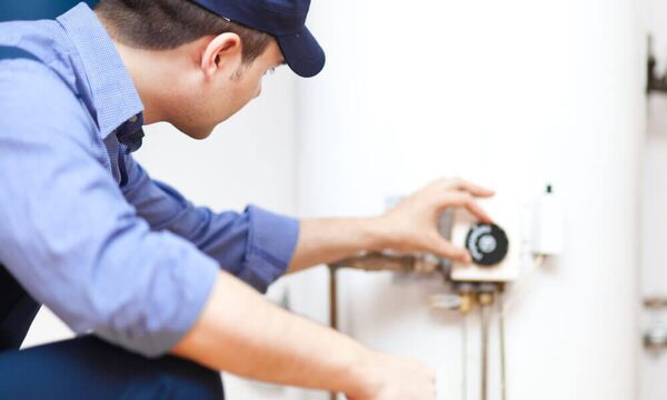 A technician in a blue shirt and cap adjusting the thermostat dial on a white hot water cylinder