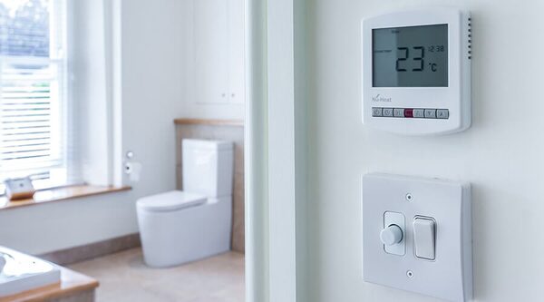 A Nu-Heat digital room thermostat set to 23°C mounted on a wall next to a light switch, with a blurred bathroom in the background