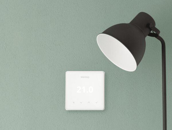 A white Warmup smart thermostat on a green wall next to a black desk lamp, displaying 21.0 degrees