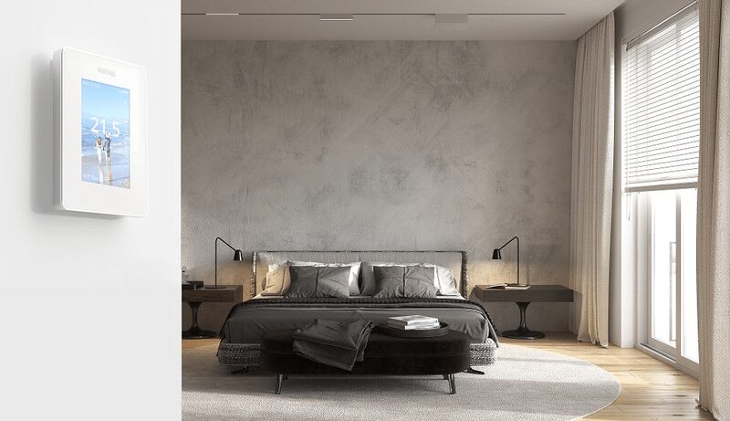 A white Warmup touchscreen underfloor heating thermostat mounted on a wall beside a modern grey bedroom