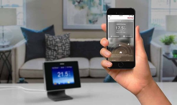 A hand holding a smartphone running the Warmup smart heating control app, with a Warmup thermostat on a stand showing 21.5°C in a living room