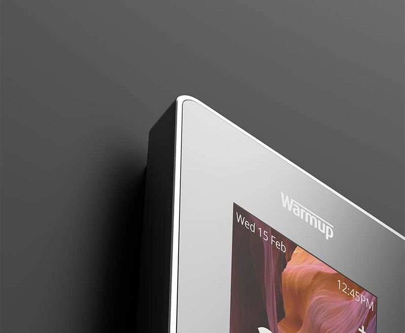 A close-up of a Warmup 6iE smart thermostat in silver mounted on a dark wall, showing part of its colour screen