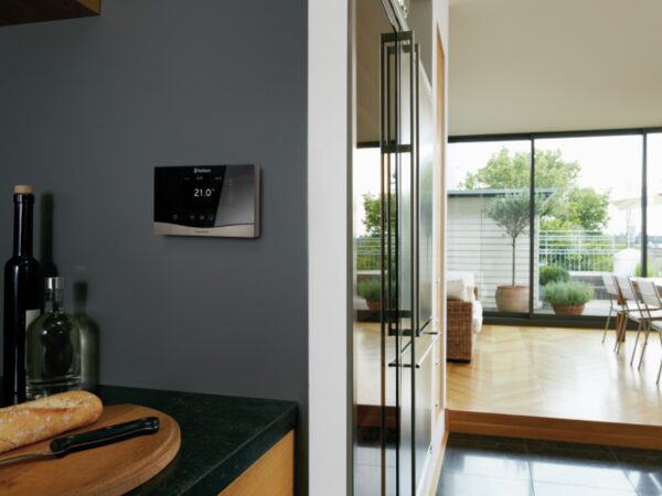 A Vaillant sensoCOMFORT smart thermostat mounted on a grey wall in a modern kitchen, showing 21.0 degrees