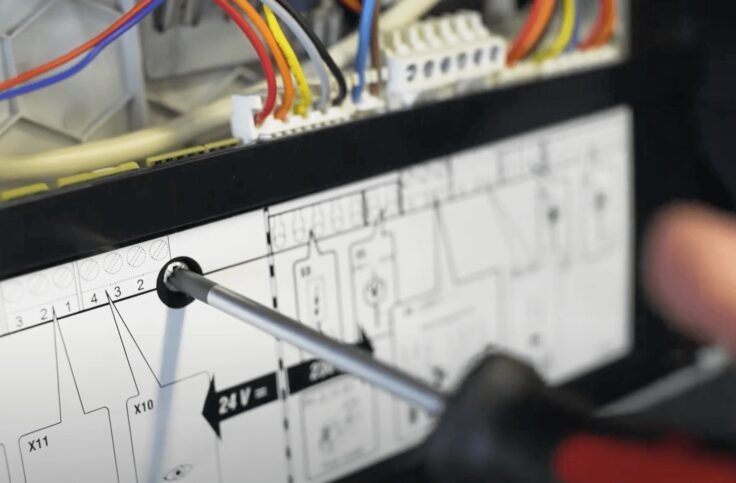 A close-up of a screwdriver adjusting wiring terminals inside a boiler's control panel with a wiring diagram label