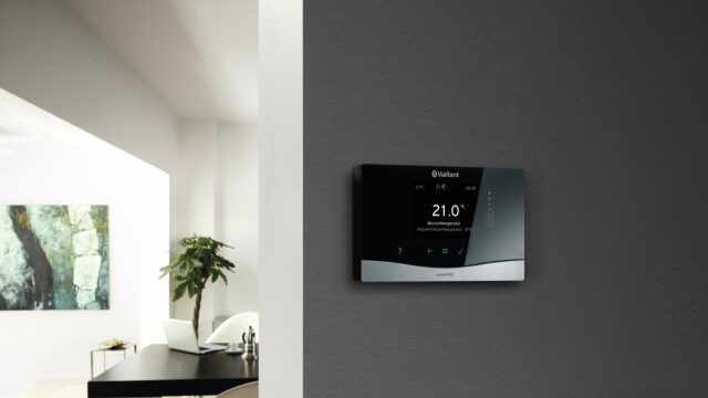 A Vaillant sensoCOMFORT wall-mounted heating control thermostat showing 21.0°C, mounted on a dark grey wall in a modern home interior